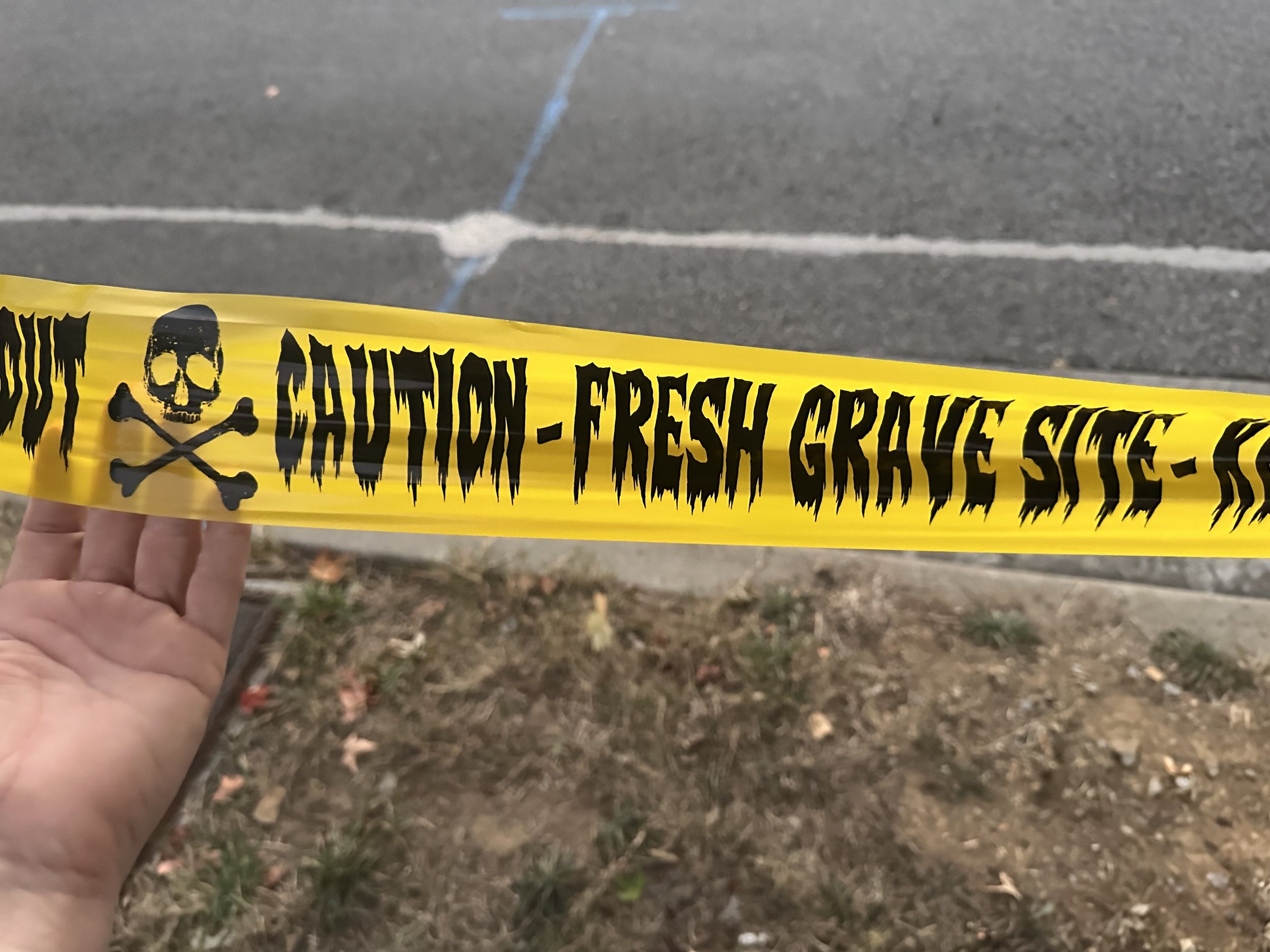 Cheap yellow caution tape surrounds a yard. The phrase, “CAUTION-FRESH GRAVE SITE” is written on it.